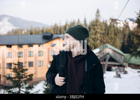 A young caucasian man enjoying an outdoor adventure in a snowy mountain setting. Dressed in warm clothing, he stands amidst trees, capturing the beaut Stock Photo