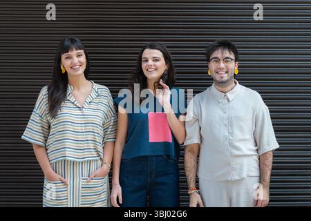 A diverse group of three friends stands together, smiling warmly at the camera. They each wear casual, trendy clothing while standing against a dark, Stock Photo