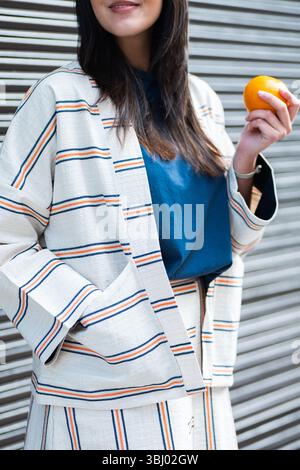 A stylish woman in a striped jacket, matching skirt, and blue top holds a vibrant orange, with a contemporary background. The scene captures a blend o Stock Photo