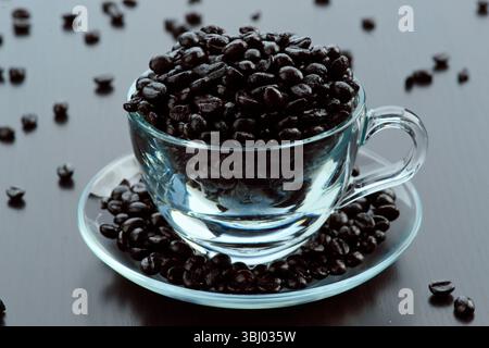A transparent glass cup filled with rich, dark coffee beans sitting on a matching saucer. Some beans are scattered on the smooth, dark surface, highli Stock Photo