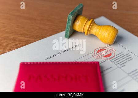 A visa application form with "APPROVED" stamp, a red passport, and a rubber stamp. International travel, permit, official consent concept. Stock Photo