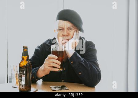 An older man sits at a table with a glass of beer, resting his head on his hand with a pensive or sad expression, appearing deep in thought Stock Photo