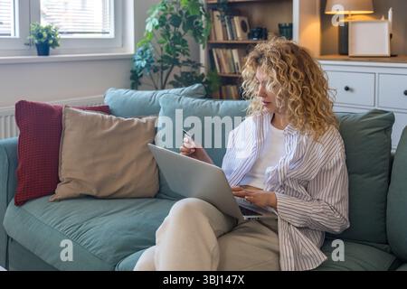 Young woman at home holding credit card and using laptop for online shopping, financial planning, booking travel, or checking banking account, relaxed Stock Photo