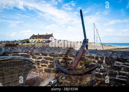 Rusty anchor displayed against a stone wall at Porlock Weir, Somerset, with colourful thatched cottages and the sea under a bright blue sky Stock Photo