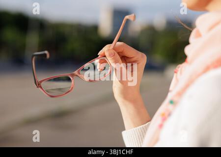 Female hand holding eyeglasses with blurred city background, representing vision care, eyesight correction, and everyday clarity. Stock Photo