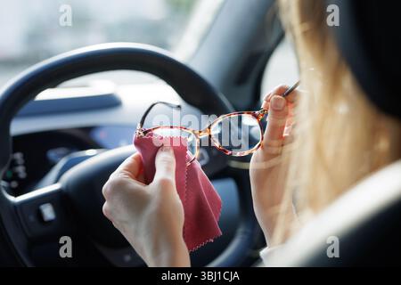 Woman cleaning glasses with microfiber cloth inside car. Focus on safe driving, hygiene, and personal care during travel. Stock Photo