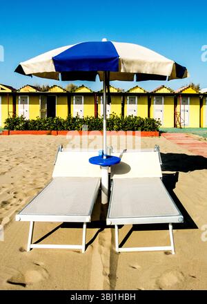 A beach scene with two lounge chairs under a blue and white umbrella. In the background, there are yellow beach huts. Stock Photo
