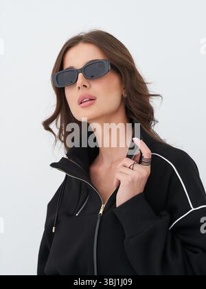 Brunette model showcases stylish black outfit with sunglasses in modern studio Stock Photo