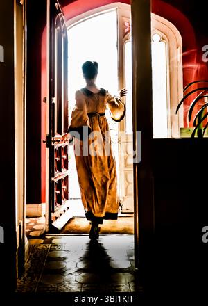 A woman in a long dress is silhouetted in a doorway, with the bright light of the outside world illuminating her from behind. Stock Photo