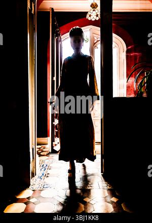 A silhouetted woman in a long dress stands in a doorway, with light streaming in from behind her. Stock Photo