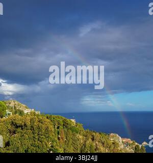 A rainbow leading from the Ionic sea over Taormina in front of a dramatic cloudy and stormy sky Stock Photo