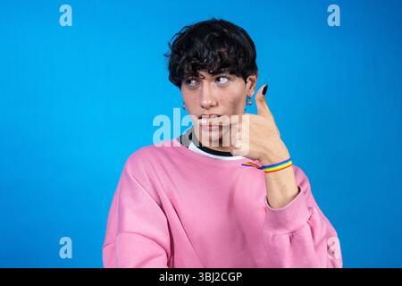 Thoughtful young man wearing pink sweatshirt and lgbt rainbow bracelet making thinking gesture on blue background Stock Photo