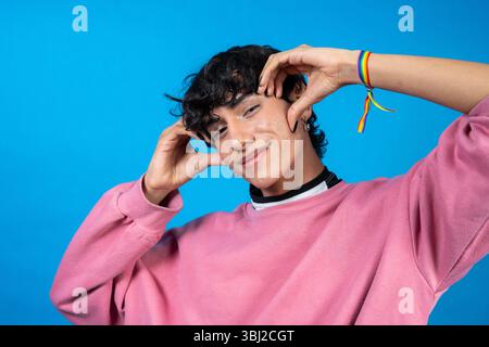 Studio portrait of a cheerful young latin man wearing a pink sweater and an lgbt rainbow bracelet, making a funny face and touching his head Stock Photo