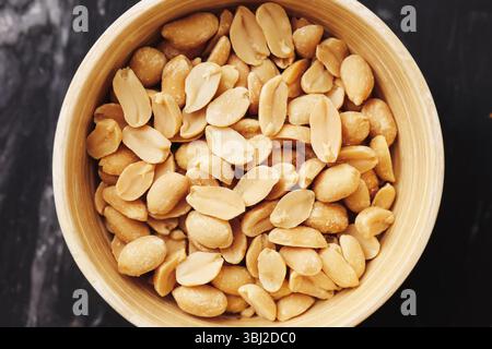 Salted peanuts in wooden bowl. Tasty crunchy snack closeup. Organic roasted nuts on marble table. Healthy appetizer rich in protein. Golden color nuts. Stock Photo