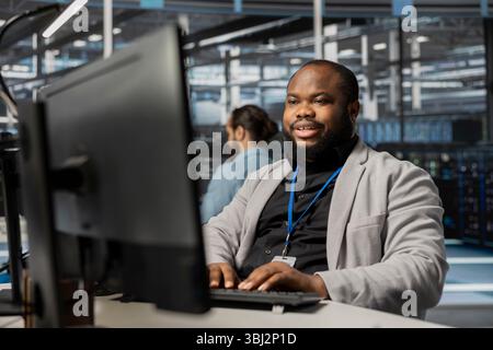 Cheerful data center employee working to update rackmounts software, configuring network virtualization. Upbeat african american man in server room verifying mainframes settings Stock Photo