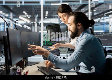 Data center manager supervising technician monitoring system performance using PC, identifying bottlenecks. Server farm teal leader overseeing worker troubleshooting glitches Stock Photo