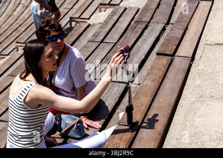 Taormina, Sizilien - Italy - 05-04-2025: Two women sitting on wooden benches in Taormina, Sicily, with one holding a phone on a tripod while taking a Stock Photo