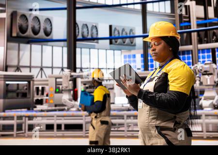 Female engineer in smart manufacturing plant monitors production line with tablet. The scene highlights industrial automation with robotic arms and solar panels, ensuring optimal factory performance. Stock Photo