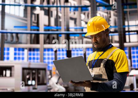 In smart manufacturing plant, black man uses laptop to oversee robotics and automation systems, ensuring optimal performance. Technician conducts quality control of industry 4.0 software in factory. Stock Photo