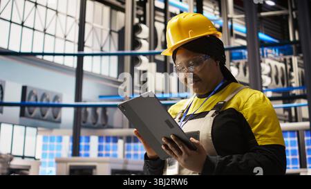 Upbeat professional working in smart factory oversees robot production lines, ensuring quality control. Industry 4.0 plant expert monitoring automated conveyor systems using notebook, camera A Stock Photo