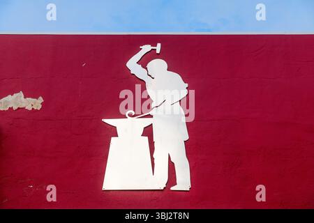 Denia, Alicante - Spain - 11-26-2024: A white metal silhouette of a blacksmith hammering on an anvil is mounted on a red wall with peeling paint, unde Stock Photo