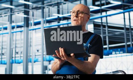 Elderly engineer uses laptop in smart factory to test automated systems, ensuring reliability. Experienced technician uses notebook to identify bottlenecks and optimize equipment, camera A Stock Photo