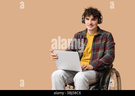 Young male programmer in headphones on wheelchair holding laptop on beige background Stock Photo