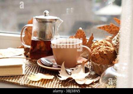 Teapot with cup of hot beverage, tasty cupcakes and autumn leaves on windowsill Stock Photo