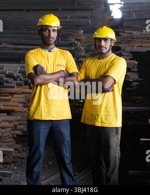 Two confident industrial workers wearing yellow safety helmets and shirts pose with arms crossed at a worksite, showcasing stregnth and teamwork. Stock Photo