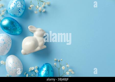 White ceramic bunny among blue and white speckled Easter eggs and delicate white flowers on a light blue background Stock Photo