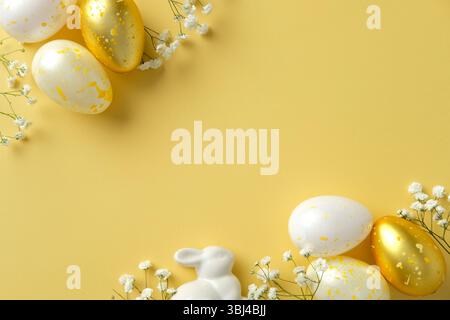 Happy Easter concept. Flat lay composition with golden speckled eggs, white ceramic bunny, and delicate flowers on a vibrant yellow background Stock Photo