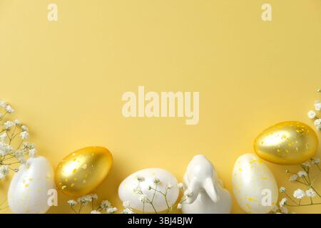 Stylish Easter flat lay. White and gold speckled eggs, a ceramic bunny figurine, and delicate white flowers on a vibrant yellow backdrop Stock Photo