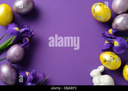 Easter flat lay composition with colorful eggs, vibrant purple irises, and a white ceramic bunny on a purple background Stock Photo
