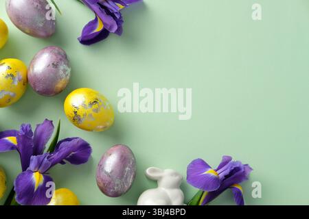Pastel Easter arrangement with decorated eggs, a white ceramic bunny, and purple irises on a green background. Stock Photo