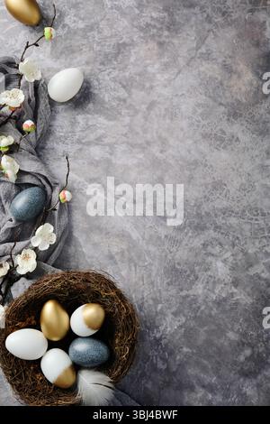 Easter flat lay with a nest of gold, white, and blue decorated eggs, blooming branches, and a grey cloth on a textured grey background Stock Photo