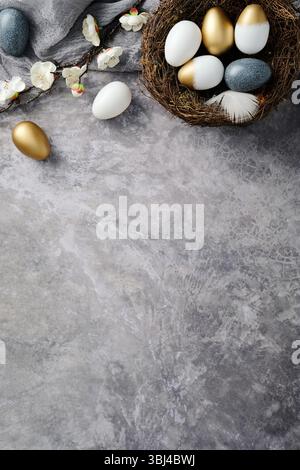 Easter flat lay composition. Top view nest with gold, white, and speckled blue eggs, surrounded by delicate white flowers on a textured gray backgroun Stock Photo