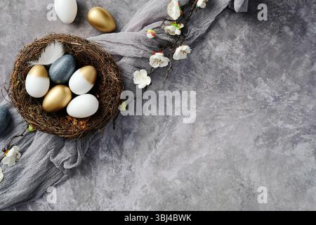 Elegant Easter flat lay composition with gold, white, and speckled eggs in a nest. Gray textured background, white cherry blossoms Stock Photo