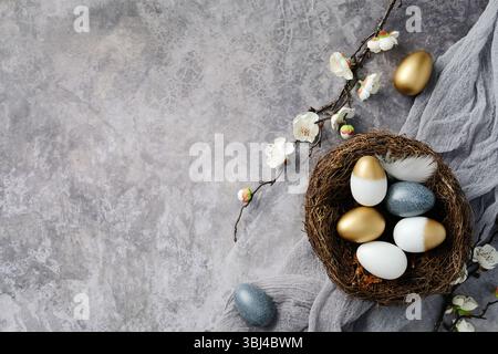 Elegant nest with Easter eggs in gold, white, and blue colors, surrounded by blooming branches on a textured gray background Stock Photo