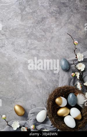 Flat lay golden and speckled Easter eggs in nest and blooming branches on textured gray background Stock Photo