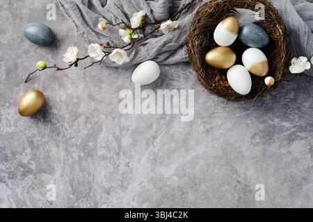 Elegant Easter flat lay composition. Nest with golden, white, and speckled eggs, surrounded by white cherry blossom branches and soft fabric on a text Stock Photo