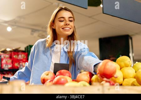 Smiling young woman in a blue shirt selecting apples at a grocery store. She reaches for a fresh red apple from a wooden fruit crate. Concept of healt Stock Photo