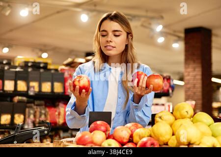 Smiling young woman in a blue shirt selecting apples at a grocery store. She reaches for a fresh red apple from a wooden fruit crate. Concept of healt Stock Photo
