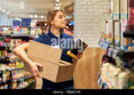Young female store worker stocking shelves in a supermarket aisle. She holds a cardboard box and a packaged item, focusing on organizing the products. Stock Photo
