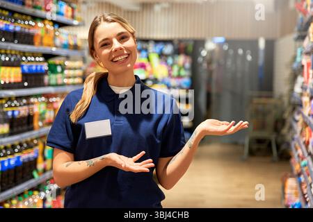 Cheerful female store worker in uniform smiling and gesturing toward supermarket shelves. Concept of customer service, helpful staff, product promotio Stock Photo