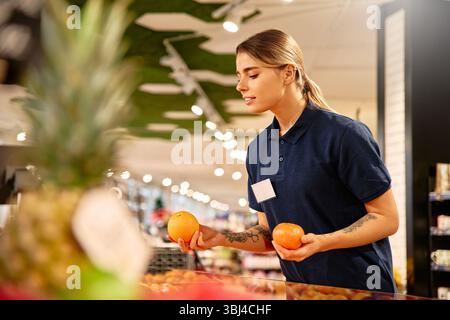 Female grocery store worker holding two oranges while inspecting the fruit display. Professional and focused employee in uniform working in produce se Stock Photo