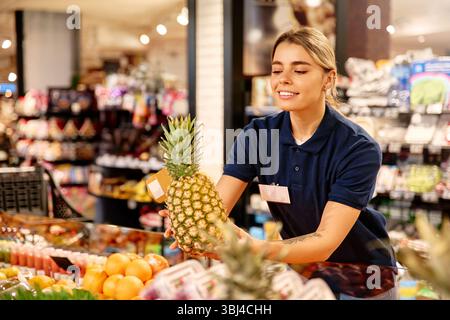 Smiling grocery store worker in uniform holding a ripe pineapple at the fresh produce section. Symbol of healthy eating and customer service in a brig Stock Photo