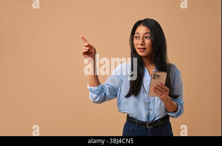 A young woman in stylish glasses holds a smartphone, gesturing as if enthusiastically explaining something important, showing strong engagement and ef Stock Photo