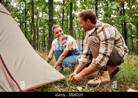 A cheerful couple happily sets up their tent in a vibrant forest, which creates a perfect and picturesque backdrop for thrilling camping adventures th Stock Photo