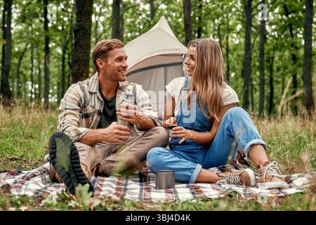 A cheerful couple sits together, relaxing on a cozy picnic blanket placed near their tent, savoring quality time in the serene embrace of nature durin Stock Photo