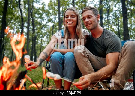 A joyful couple roasting delicious marshmallows over a warm campfire in the serene woods, relishing in their quality time spent together while embraci Stock Photo